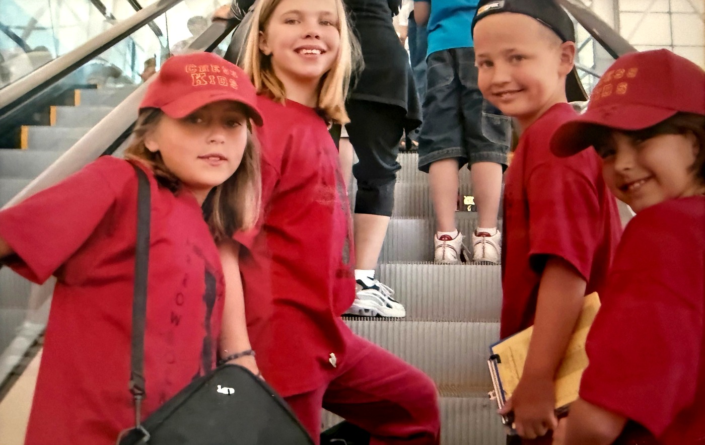 Four children in matching red chess team caps and shirts riding an escalator into a tournament venue, one carrying a board bag, another a folder and notation pad.