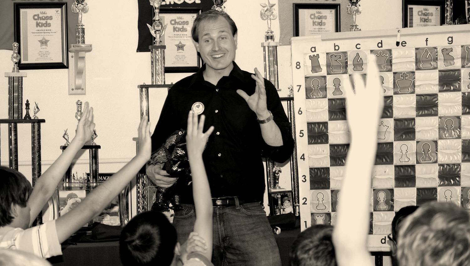 Daniel Vellotti mid-sentence in front of a demonstration board, a plush dragon under one arm, with every child in the room holding a hand up.
